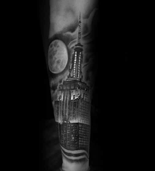 Empire State Building tattoo illuminated under a full moon, featuring dramatic shadows and detailed black shading on a man's forearm.
