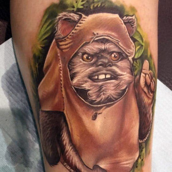 Lifelike Ewok tattoo with a tan hood, playful grin, and raised finger, surrounded by lush green forest detailing.