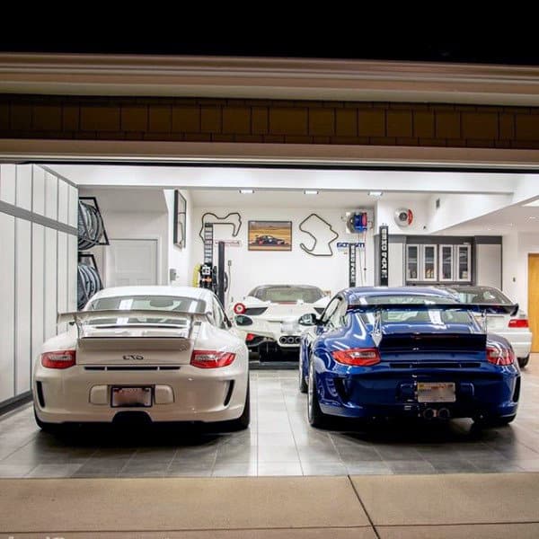A sleek, well-lit home garage featuring white and blue Porsches alongside a Ferrari, with racing memorabilia adorning the walls.