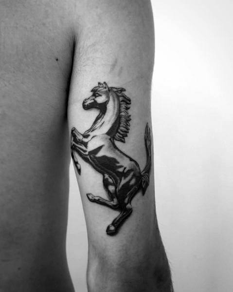 Black-and-grey Ferrari prancing horse tattoo on man's upper arm, symbolizing power, freedom, and automotive legacy.