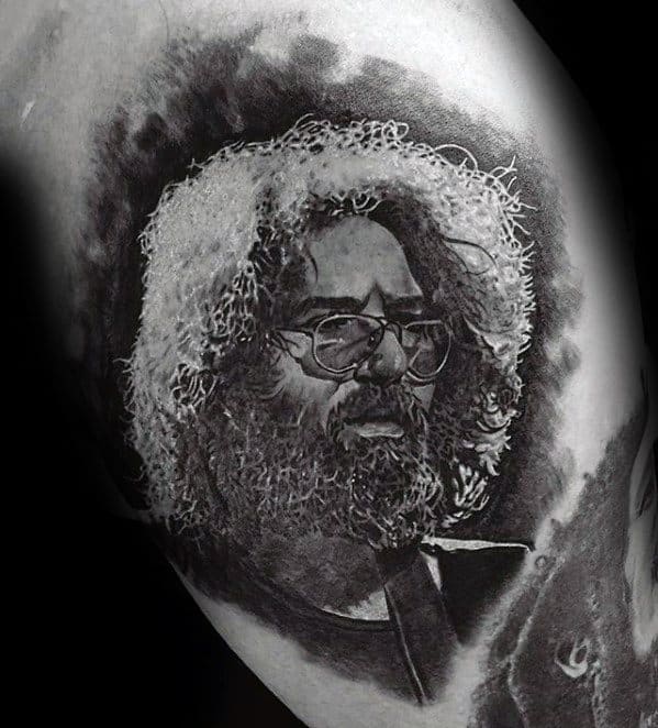 Realistic black and grey tattoo portrait of Jerry Garcia, featuring detailed hair, glasses, and a contemplative expression.