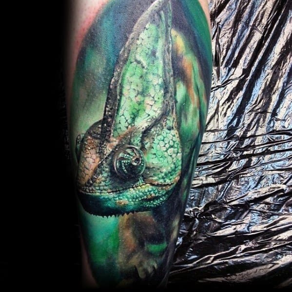 Hyper-realistic chameleon tattoo with vivid green tones, detailed skin texture, and a soft, abstract background blending into the arm