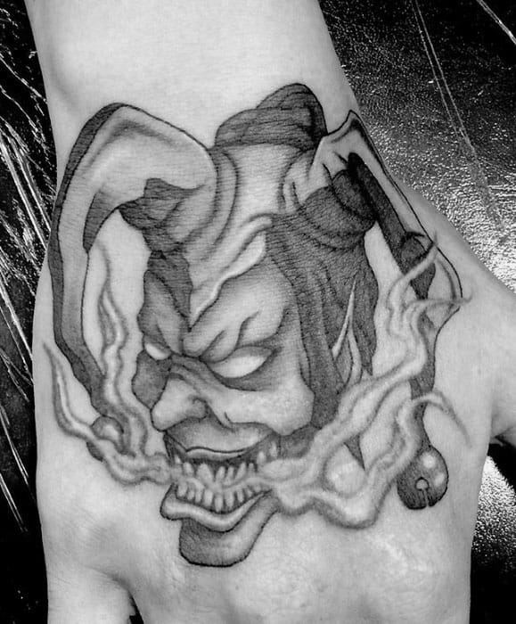 A black and gray tattoo of a jester's face with a skull-like expression, surrounded by wisps of smoke, placed on the back of the hand.