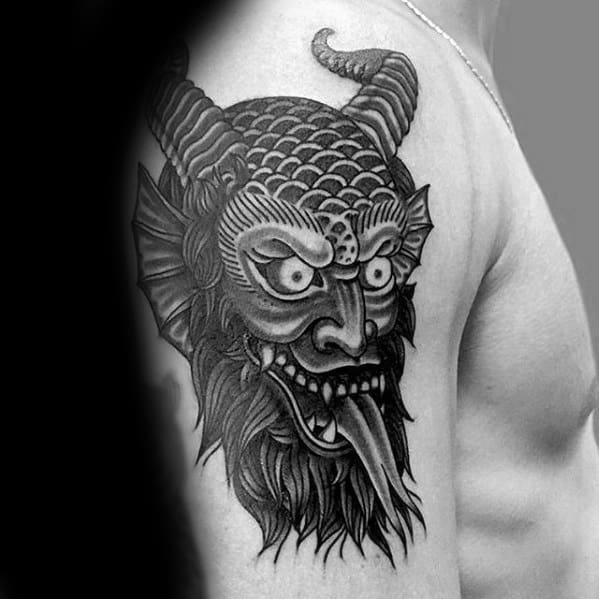 Black and grey Krampus tattoo on upper arm showing a fierce horned face with scale-like texture, wild eyes, fangs, and a long tongue.