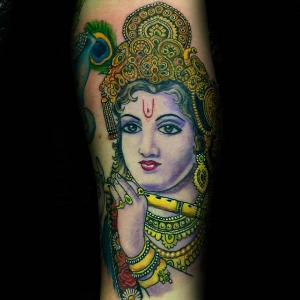 Colorful Krishna tattoo showing his serene face with a yellow flute, detailed jewelry, and a peacock feather near the crown.