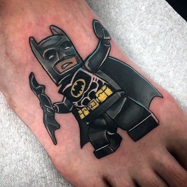 Colorful tattoo of Lego Batman on the foot, featuring his black suit, yellow utility belt, and batarang in a dynamic pose.