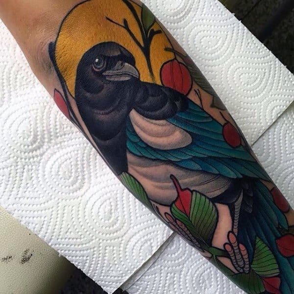 Colorful magpie tattoo on forearm, featuring bold blue and black feathers, red berries, and a yellow backdrop for a striking, artistic look.
