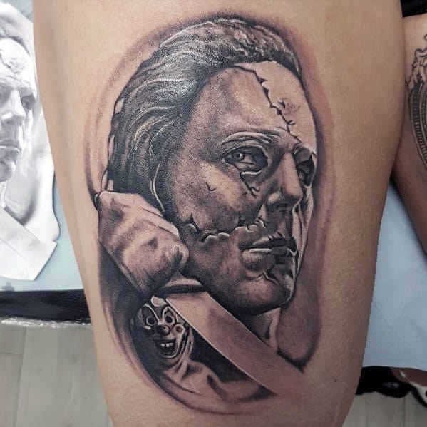 Black and gray Michael Myers tattoo with a cracked mask, gripping a knife, and a haunting small clown figure in the blade's reflection.