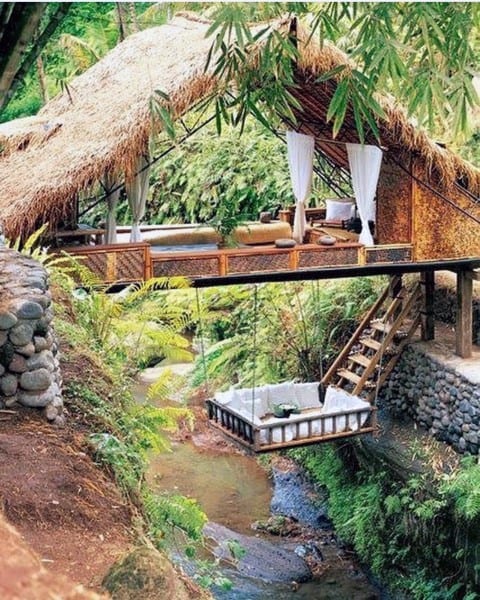 Rustic hanging bed with cushions suspended over a creek under a tropical thatched-roof retreat.