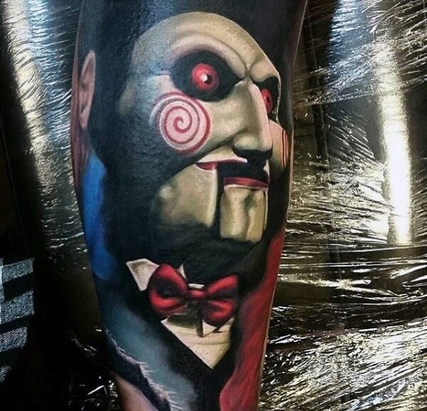 Realistic color tattoo of Billy the Puppet from *Saw*, featuring his red spiral cheeks, glowing eyes, and detailed shading for a chilling horror effect.