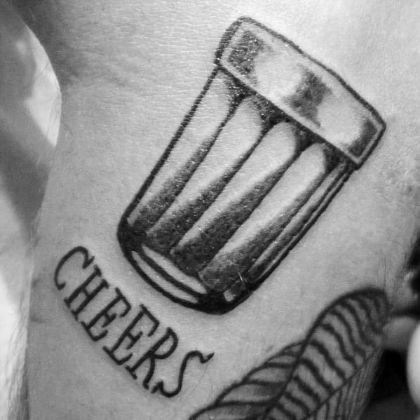 Black and grey shot glass tattoo with bold shading, tilted for a dynamic effect, accompanied by the word "CHEERS" in a stylish font.