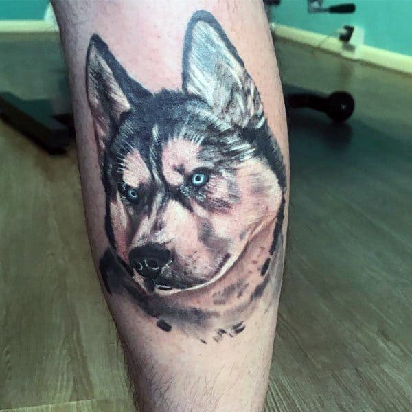 Realistic Siberian Husky tattoo on a man's leg, featuring piercing blue eyes, detailed fur shading, and a strong, lifelike expression.