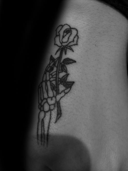 Side hand tattoo for men featuring a skeletal hand holding a rose, combining dark and delicate elements in a bold line design.