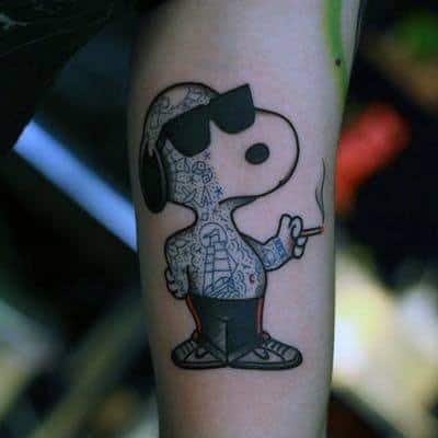 Tattoo of Snoopy wearing sunglasses, sneakers, and covered in intricate tattoos, smoking a cigarette, placed on a person's arm.