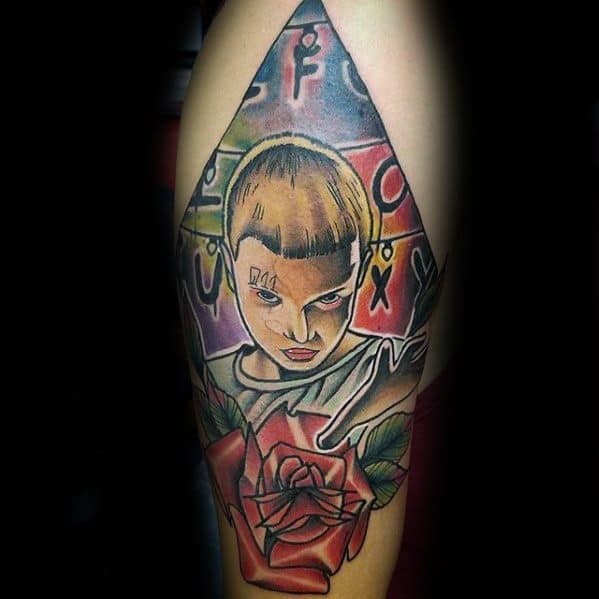 Colorful tattoo of Eleven with an intense gaze, standing before the alphabet wall, framed by a red rose and triangle shape.