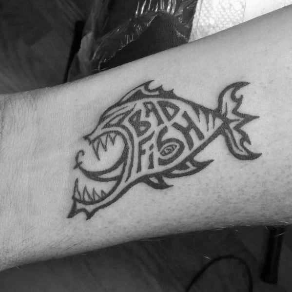 Black ink tattoo of a stylized fish with sharp teeth, featuring the words “Bad Fish” creatively integrated into its body.