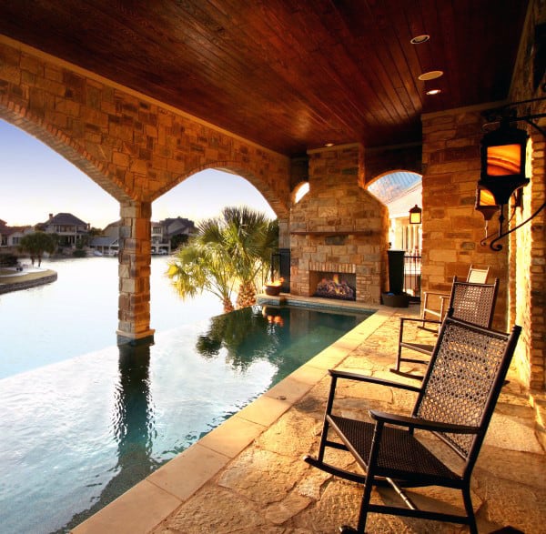 Covered patio with stone arches, infinity pool, rocking chairs, and fireplace overlooking a tranquil lake at sunset