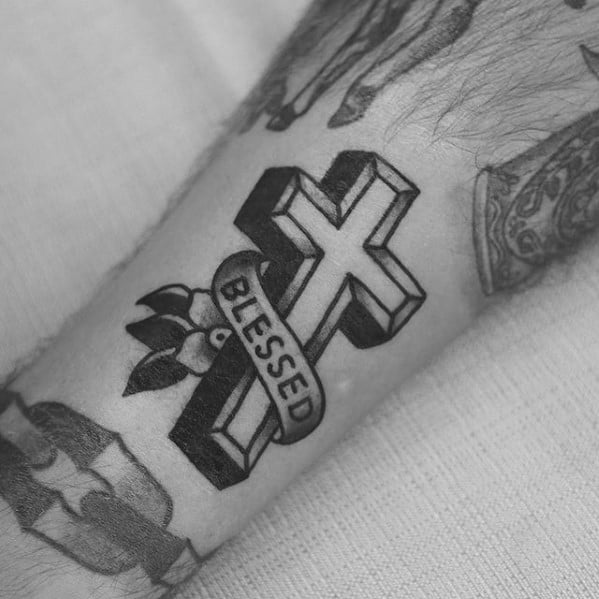Black 3D cross tattoo with a "Blessed" banner wrapped around it and leaves in the background, inked on the forearm.