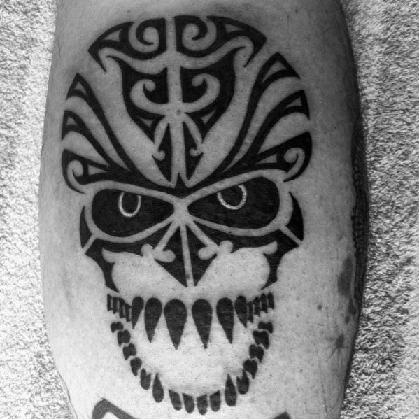 Fierce tribal skull tattoo with bold black shapes, intense eyes, and sharp teeth forming a striking symmetrical design on the leg.