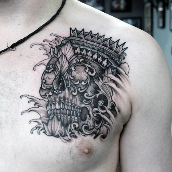 Black and grey Tibetan skull tattoo on the chest with ornamental crown, flowing smoke, and intricate floral-style details.