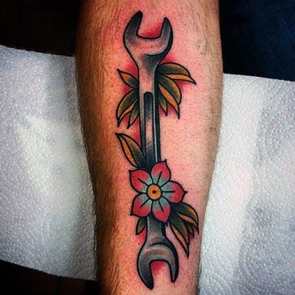 Colorful tattoo of a wrench with surrounding green leaves and a red flower in a bold traditional style on the forearm.