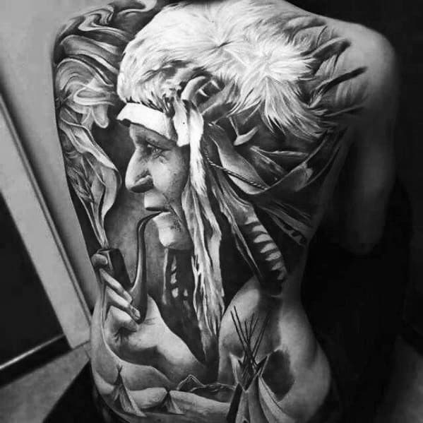 Large full-back tattoo in black and grey featuring a realistic Native American chief wearing a feathered headdress, smoking a pipe, with teepees below.