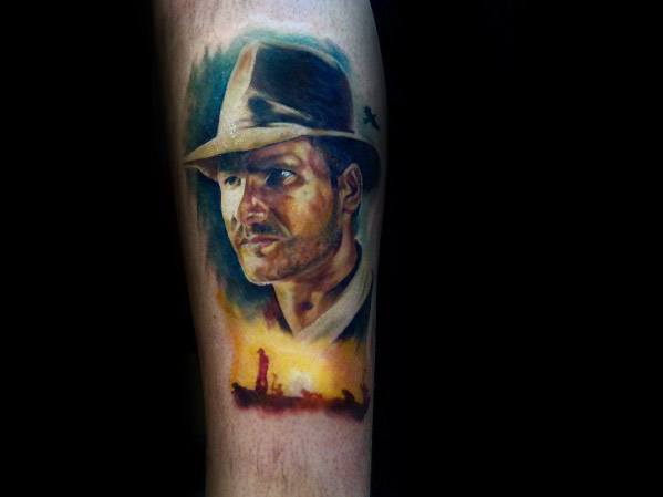 Vivid Indiana Jones tattoo featuring his iconic fedora and determined expression, with a desert adventure scene glowing below.