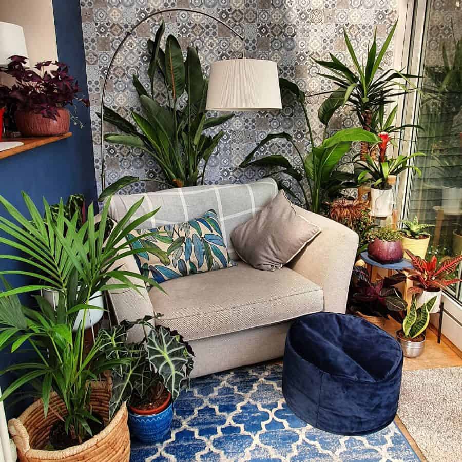 Cozy nook with a beige armchair, leafy plants, patterned wallpaper, and blue geometric rug illuminated by a standing lamp