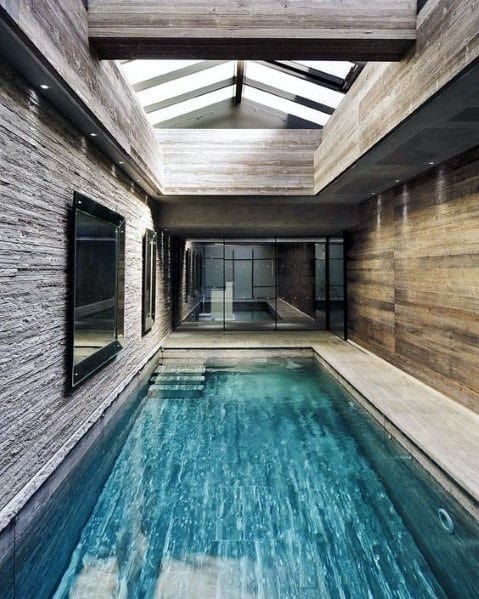 Modern indoor pool with textured stone walls and elegant blue tiles, illuminated by natural light through a skylight.