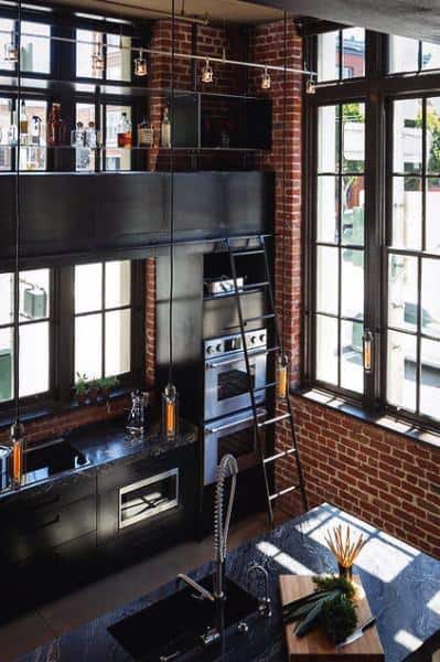 Industrial kitchen with brick walls, tall windows, and black cabinetry with a ladder.