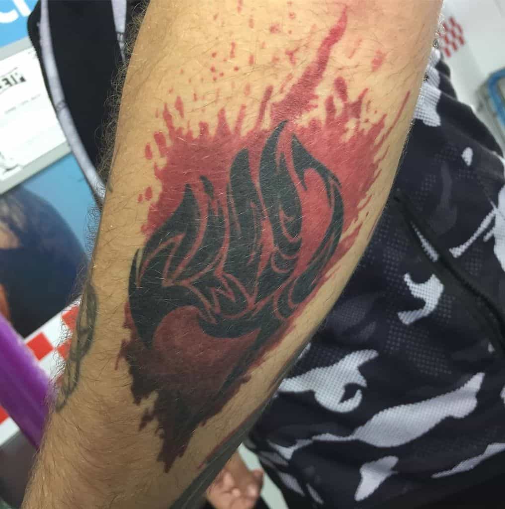 Bold Fairy Tail guild emblem tattoo with a dark, fiery design, surrounded by a striking red splash effect on the forearm.