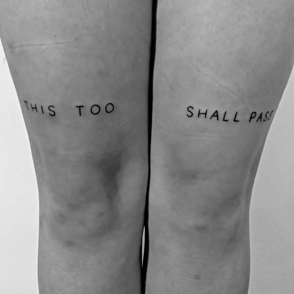 A "This Too Shall Pass" tattoo on each thigh, with the words split across both legs. The image is in black and white.
