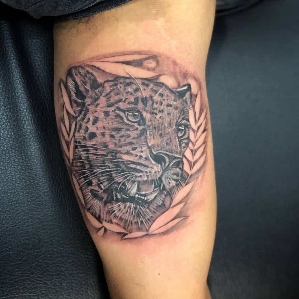 A black and grey tattoo of a jaguar's face framed by stylized leaves, showcasing intricate shading and detailing, placed on the upper arm.