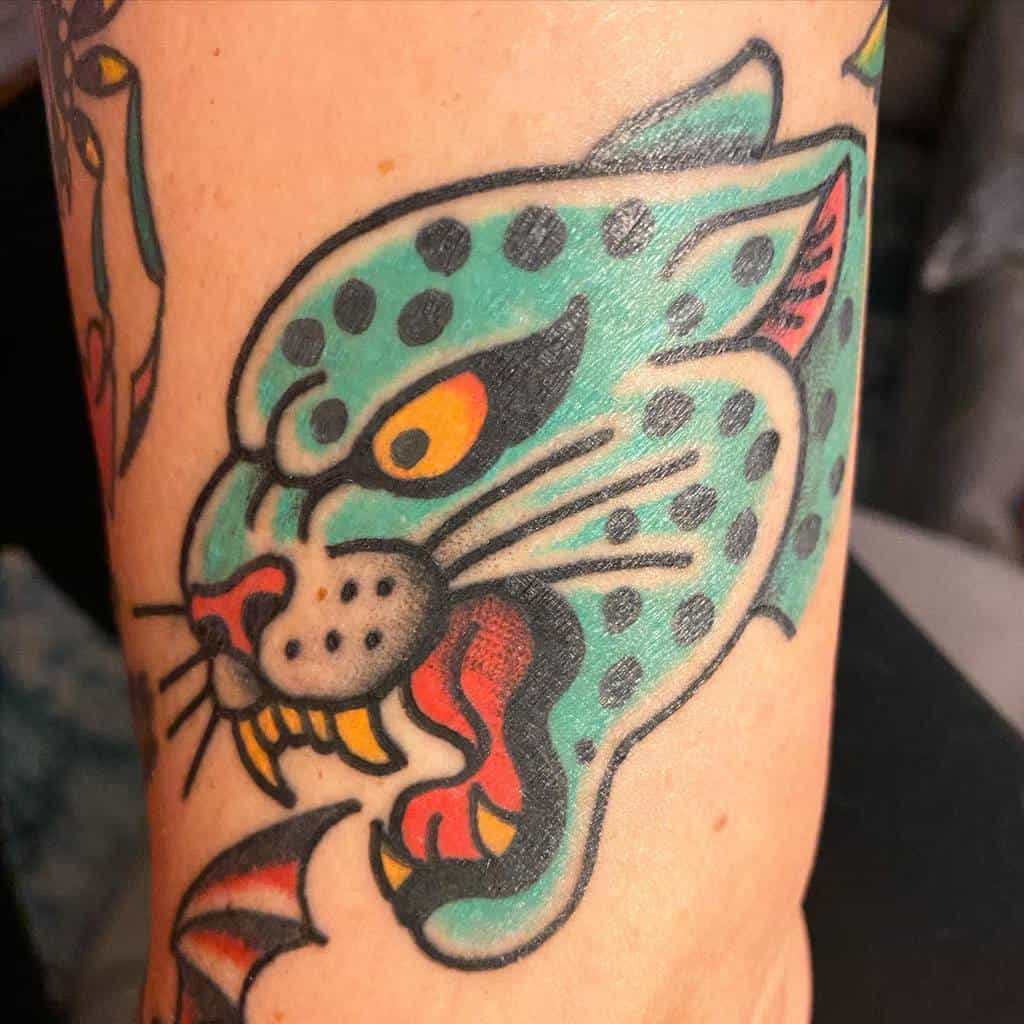 A bold traditional-style tattoo of a snarling jaguar, featuring vibrant turquoise and yellow colors, with black outlines and sharp, expressive details on the arm.