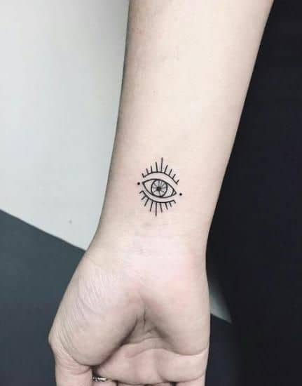 Small minimalist tattoo of an all-seeing eye with radiating lines, inked in fine black lines on the wrist.
