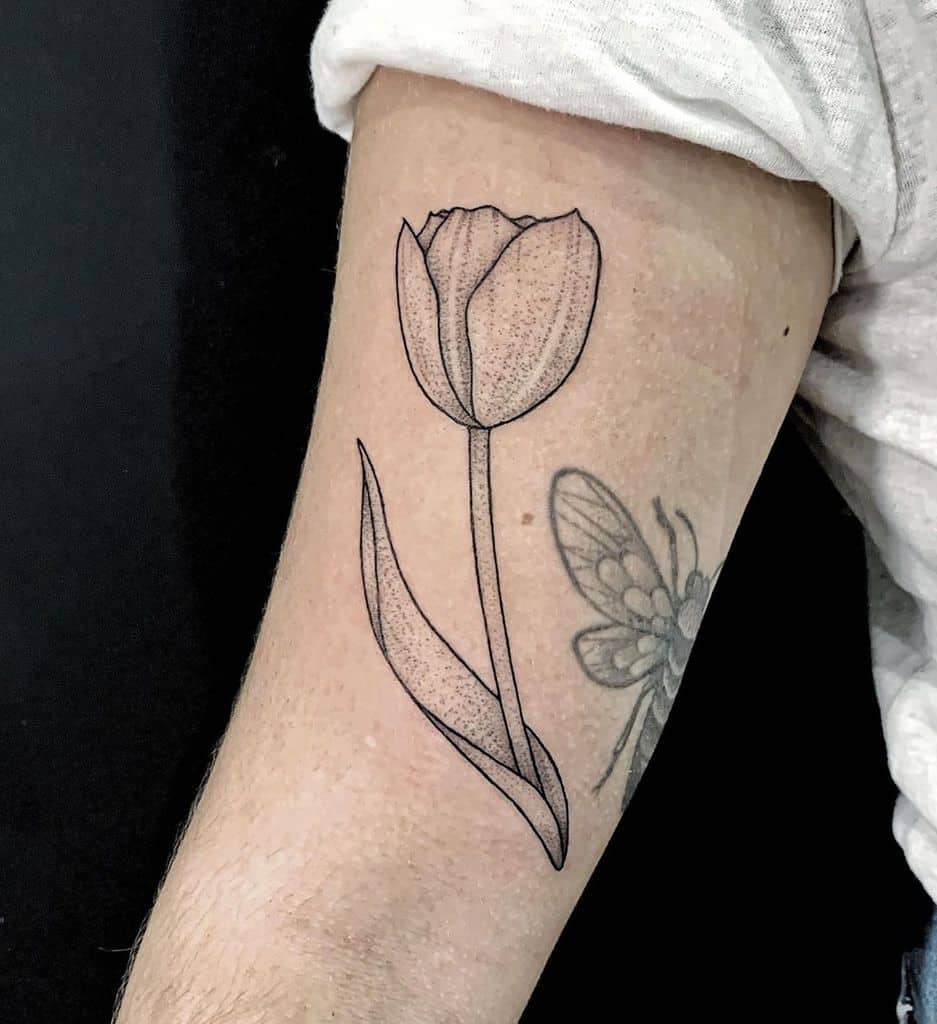 A black-line tulip tattoo on the upper arm, featuring fine dotwork shading and a minimalist design for an elegant floral look.