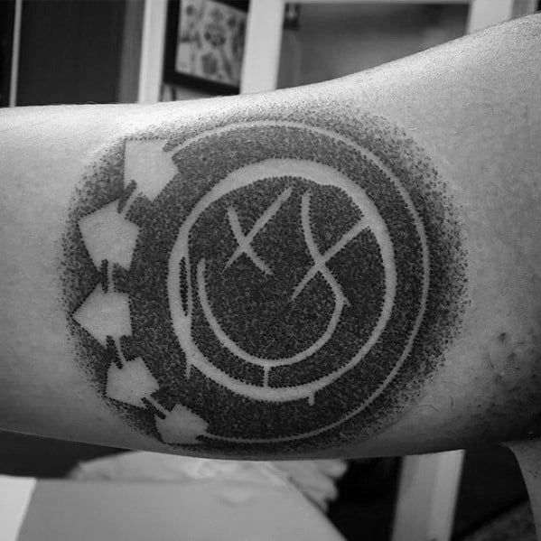 Black and gray tattoo of the Blink-182 smiley face logo with shading, giving it a soft, dotted effect around the edges, placed on the forearm.