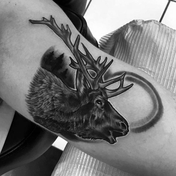 Realistic black and grey elk tattoo on the bicep, featuring intricate shading, bold antlers, and a circular design for a dynamic look.