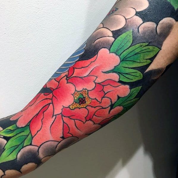 A bold inner-arm bicep tattoo featuring a vibrant Japanese flower design, surrounded by soft cloud-like shading and striking green leaves