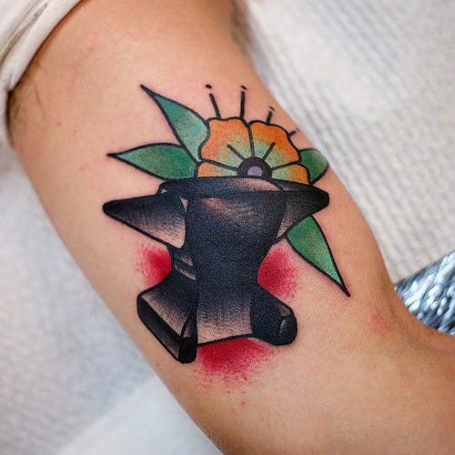 Traditional anvil tattoo with bold black shading, set against a vibrant orange flower and green leaves, with red background accents.