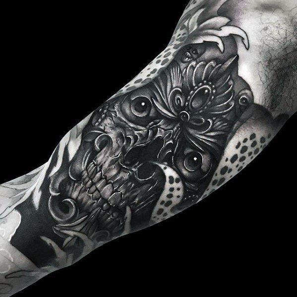 Black and grey Tibetan skull tattoo on the inner arm with deep shading, wide eyes, and ornate crown-like detailing.