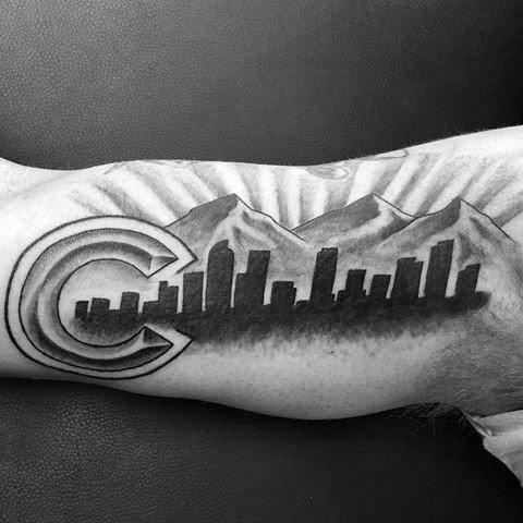 Black and grey Denver skyline tattoo on inner arm with mountains and a bold Colorado “C” symbol blending into the cityscape.