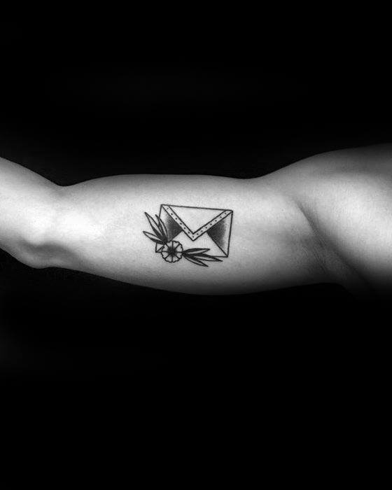Tattoo of an envelope with flowers and leaves on a flexed bicep against a black background