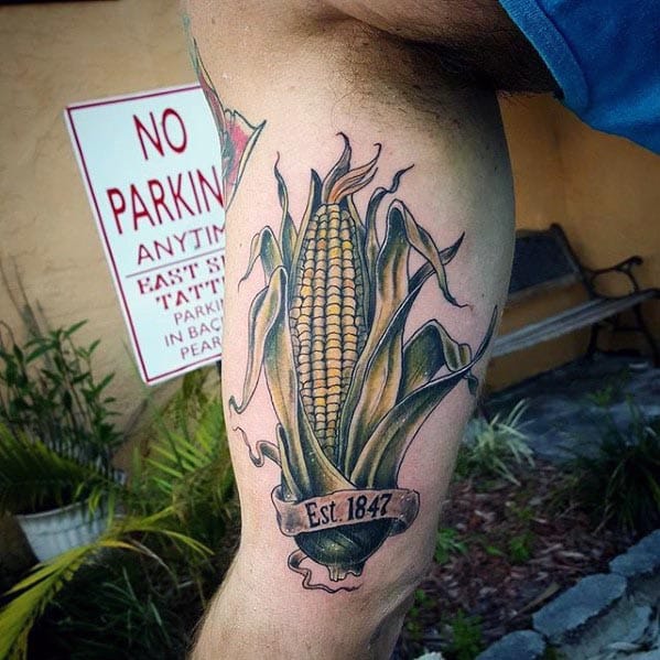 Detailed tattoo of a corn cob with husks and a banner reading 'Est. 1847,' featuring realistic shading and earthy tones.