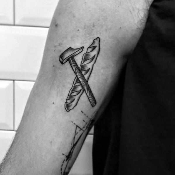 Black ink tattoo of a crossed hammer and baguette, symbolizing strength and craftsmanship, with fine shading for a bold yet minimalist look.
