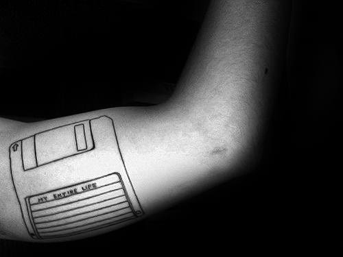 Simple black line tattoo of a floppy disk on the inner arm with the text “My Entire Life” written on the label.