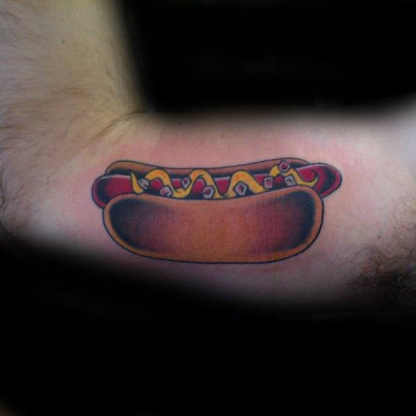 Hot dog tattoo featuring a sausage with mustard, ketchup, and onions in a bun on the upper arm.