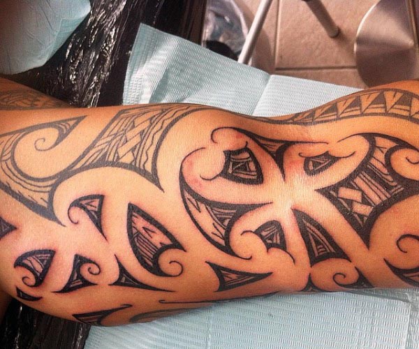 Bold Polynesian tribal tattoo covering the inner bicep, featuring intricate black linework, curves, and geometric patterns