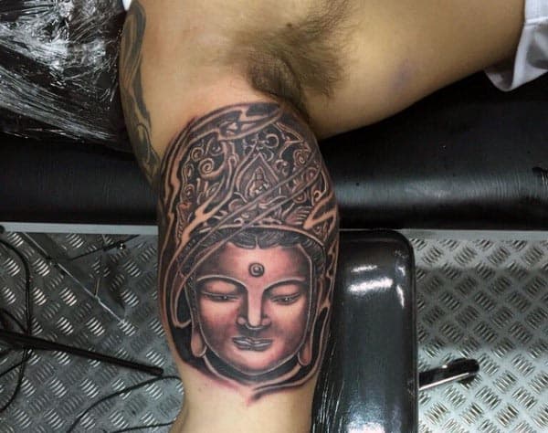 Black and grey Buddha tattoo on the inner bicep, featuring intricate headdress details, smooth shading, and a peaceful facial expression