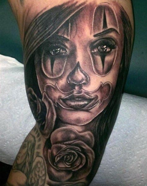 Black and grey tattoo of a woman with clown makeup, featuring realistic shading, intense eyes, and roses on the inner bicep